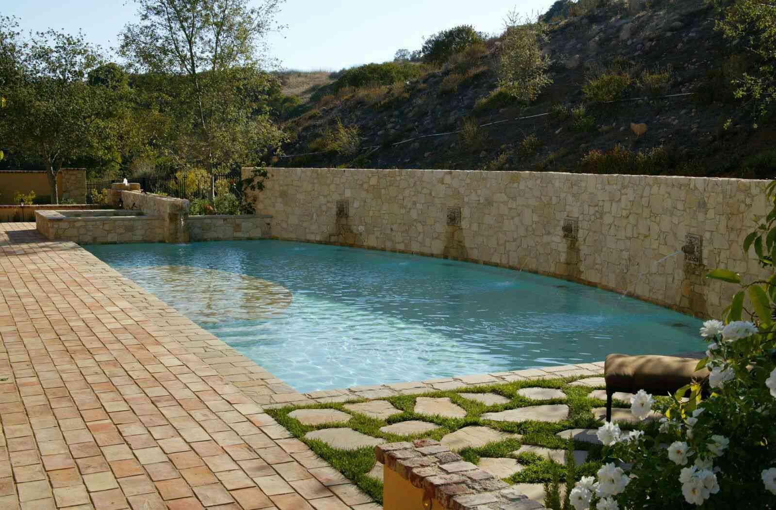 Piscine méditerranéenne avec un mur en carreaux beiges et une allée de pierre herbeuse.